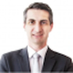 Bu, profesyonel siyah takım elbise giyen bir adamın resmi. Beyaz yakalı bir gömleği ve siyah bir kravatı var. Saçları kısa ve düzgünce taranmış, yüzü ise sinekkaydı tıraş edilmiş. Beyaz bir duvarın önünde duruyor, kollarını önünde kavuşturmuş ve yüzünde ciddi bir ifade var. Duruşu kendinden emin ve güçlüdür. Adamın koyu renk gözleri var ve doğrudan kameraya bakıyor. Yeteneklerine güvenen, profesyonel ve güçlü bir adam gibi görünüyor. Önüne çıkacak her türlü zorluğu göğüslemeye hazır görünüyor.