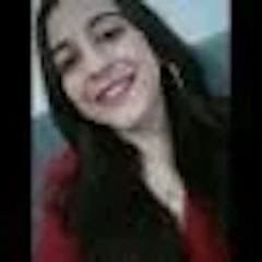 Genç kadın telefonuyla selfie çekerken ışıl ışıl gülümsüyor. Dalgalı kahverengi saçları yüzünden geriye doğru toplanmış. Açık pembe bir gömlek giyiyor ve boynunda gümüş bir kolye var. Gözleri kapalı ve yüzünde huzurlu bir ifade var. Beyaz bir duvarın önünde duruyor, kolunu uzatmış, elinde telefon var. Bu anda memnun ve rahat görünüyor ve gülümsemesi odayı aydınlatıyor.