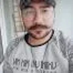 Bıyıklı ve sakallı genç bir adam selfie çekiyor. Telefonunu önünde tutuyor ve kararlı bir ifadeyle objektife bakıyor. Yüzündeki kıllar düzgün, kalın bir bıyık ve dolgun, kalın bir sakal şeklinde bakımlı. Siyah bir tişört ve koyu renk bir kot pantolon giymiş. Arka plan net değil ama kadrajda bir oturma odasının izleri var. Bu selfie'yi çekerken gözleri odaklanmış ve konsantre olmuş durumda. Anın tadını çıkarıyor gibi görünüyor ve yüzü sıcak, mutlu bir ifadeyle aydınlanıyor.
