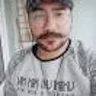 Bıyıklı ve sakallı genç bir adam selfie çekiyor. Telefonunu önünde tutuyor ve kararlı bir ifadeyle objektife bakıyor. Yüzündeki kıllar düzgün, kalın bir bıyık ve dolgun, kalın bir sakal şeklinde bakımlı. Siyah bir tişört ve koyu renk bir kot pantolon giymiş. Arka plan net değil ama kadrajda bir oturma odasının izleri var. Bu selfie'yi çekerken gözleri odaklanmış ve konsantre olmuş durumda. Anın tadını çıkarıyor gibi görünüyor ve yüzü sıcak, mutlu bir ifadeyle aydınlanıyor.