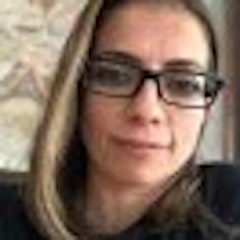 Resimdeki kadının omuzlarına dökülen ve yüzünü çerçeveleyen uzun, dalgalı kahverengi saçları var. Koyu kahverengi gözlerini vurgulayan gümüş çerçeveli gözlükler takıyor. Yüzünde rahat bir ifade ve kendine güvenen bir hava var. Beyaz, uzun kollu bir gömlek giyiyor ve üstteki birkaç düğmesi açık, göğsünü ortaya çıkarıyor. Boynunda sade, gümüş bir kolye ve ona uygun küpeler var. Dudakları sanki bir şey söylemek üzereymiş gibi hafifçe ayrık. Etrafını saran nazik, kibar bir aurası var ve varlığı sakinleştirici.