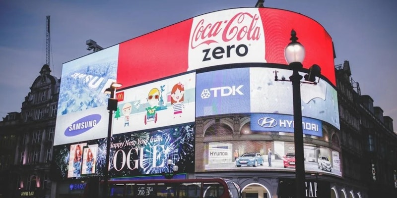 Resimde bir bina üzerindeki büyük bir reklam panosu görülüyor. Üst ortada mavi zemin üzerine beyaz bir harf ve sol alt köşede bir logonun yakın çekimi yer alıyor. Logonun sağında, üzerinde metin bulunan bir tabelanın yakın çekimi yer alıyor. Daha sağda, üzerinde bulanık bir insan başı resmi olan başka bir tabelanın yakın çekimi yer alıyor. Reklam panosu reklamlar içeriyor ve parlak bir şekilde aydınlatılmış. Reklam panosunun arka planı açık sarı-kahverengi renktedir.