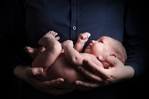 Bir kişi kucağında bir bebek tutmaktadır ve bebeğin başı omzuna yaslanmıştır. Bebeğin yüzü görünüyor ve kameraya bakıyor, gözleri kapalı ve yüzünde memnun bir ifade var. Bebeğin elleri yumruk halindedir ve elleri kişinin gömleğini kavrarken görülebilir. Kişinin elleri bebeği sıkıca kavramış, güvenli ve rahatlatıcı bir kucaklama sağlıyor. Bebeğin ayakları da görülebiliyor, ayak parmakları hafifçe kıvrılmış. Arka planda mavi bir kumaşın yakın çekimi görülüyor.
