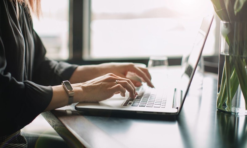 Bir kişi bir dizüstü bilgisayarda çılgınca yazıyor. Dizüstü bilgisayar beyaz arka planlı bir masanın üzerinde durmaktadır. Kişi kısa saçlı ve gözlük takıyor. Elleri klavyenin üzerindedir ve yüz ifadesi odaklanmış ve yoğundur. Dizüstü bilgisayarın ekranında, karmaşık detaylara sahip siyah beyaz bir saat görüntüsü vardır. Dizüstü bilgisayarın sağında cam bir kavanoz içinde küçük bir bitki görülmektedir. Kişinin sağ elinin orta parmağında basit bir altın yüzük görülüyor. Görüntüde hafif bir bulanıklık var ve bu da genel olarak hareket ve hız hissi veriyor.