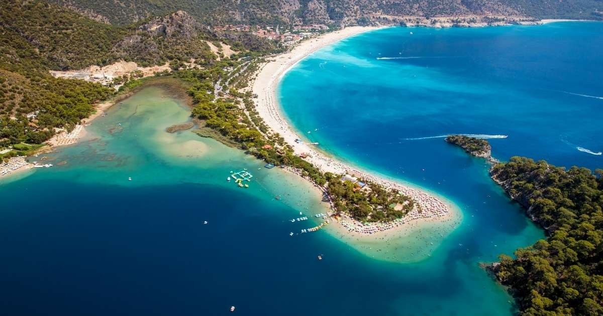 Bu, pırıl pırıl mavi bir su kütlesine doğru uzanan uzun bir kum şeridine sahip bir plajın havadan görünümüdür. Kıyı boyunca dağılmış, bazıları yürüyen, bazıları da plaj sandalyelerine uzanmış insanlar görülüyor. Sahilin ilerisinde, güneşten biraz gölge sağlayan binalar ve ağaçlar var. Uzakta, pitoresk bir fon sağlayan bir orman görülebilir. Gökyüzü parlak mavi ve güneş ışıl ışıl parlayarak pastoral bir atmosfer yaratıyor.