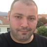 Siyah saçlı ve açık mavi tişörtlü bir adam beyaz bir duvarın önünde duruyor ve sağ elinde bir akıllı telefon tutuyor. Sol elini önüne doğru uzatmış ve telefonunun ön kamerasına bakarak selfie çekiyor. Yüzünde parlak bir gülümseme var ve anın tadını çıkarıyor gibi görünüyor. Işık yumuşak ve görüntüdeki renkler canlı. Adam dik bir duruş sergiliyor ve kameraya kendinden emin bir şekilde bakıyor.