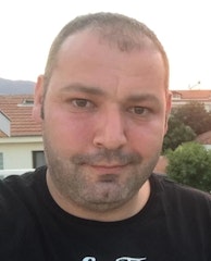 Siyah saçlı ve açık mavi tişörtlü bir adam beyaz bir duvarın önünde duruyor ve sağ elinde bir akıllı telefon tutuyor. Sol elini önüne doğru uzatmış ve telefonunun ön kamerasına bakarak selfie çekiyor. Yüzünde parlak bir gülümseme var ve anın tadını çıkarıyor gibi görünüyor. Işık yumuşak ve görüntüdeki renkler canlı. Adam dik bir duruş sergiliyor ve kameraya kendinden emin bir şekilde bakıyor.