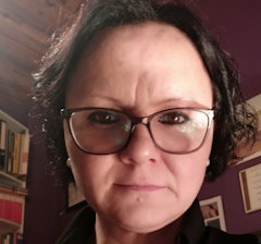 Genç bir kadın açık renkli bir duvarın önünde duruyor, mavi bir ceket, kot pantolon ve beyaz spor ayakkabı giyiyor. Bir elinde akıllı telefonunu tutuyor ve diğer eliyle selfie çekiyor. Yüz ifadesi neşeli ve kendinden emin, dudaklarında hafif bir gülümseme var. Kısa kahverengi saçları var ve gözleri makyajla vurgulanmış. Bir kalçası öne doğru itilmiş şekilde duruyor ve bacakları hafifçe ayrık. Işık yumuşak ve doğal, arka plan ise nötr.