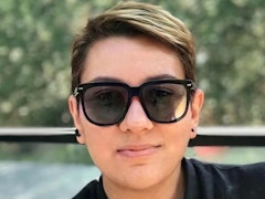 Bu, koyu renkli güneş gözlüğü takan bir kişinin görüntüsüdür. Kişi uzun, koyu renk saçlara sahip ve açık havada bir kafede oturuyor. Kollarını kavuşturmuş ve yüzünde hafif bir gülümseme var. Kişi siyah bir tişört ve mavi kot pantolon giymektedir ve güneş gözlükleri klasik havacı tarzındadır. Arkalarındaki gökyüzü açık ve parlak ve ışık güneş gözlüklerinin camlarından yansıyor. Kişi kendinden emin, rahat ve günün tadını çıkarmaya hazır görünüyor.