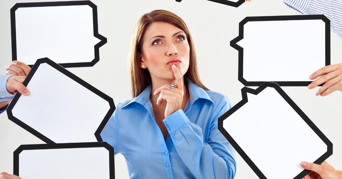 A woman wearing a light blue, collared shirt is standing in front of a white background. She is looking to the right side and has her right hand resting on her chin. Her left hand is outstretched, and a black ring can be seen on the ring finger. Her left hand is also holding a white sign. In the background, an additional hand is visible, holding a black object. The woman has a neutral expression and her hair is styled in a neat, short cut. Her shirt has two pockets on the front, and the sleeves are rolled up to her elbows.