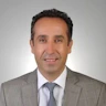 Koyu renk takım elbiseli ve açık mavi kravatlı orta yaşlı bir adam beyaz bir arka planın önünde duruyor. Ellerini önünde kavuşturmuş, dümdüz karşıya bakıyor. Saçları düzgünce taranmıştır ve bir gözlük takmaktadır. Yüzünde kendinden emin bir ifade var ve gözleri odaklanmış. Profesyonel ve başarılı görünüyor. Beden dili, önüne çıkan her türlü zorluğu üstlenmeye hazır olduğunu gösteriyor.