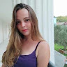 Bir kadın aydınlık bir dış mekânda kameranın önünde durmaktadır. Üzerinde parlak desenli beyaz bir üst ve koyu renk bir kot pantolon var. Saçları dağınık bir topuz yapılmış ve yüzü hafifçe yana çevrilerek yumuşak yüz hatları ortaya çıkarılmış. Gülümsüyor ve ifadesi neşe ve güven dolu. Elleri kalçalarında, duruşu kendinden emin ve rahat. Kameraya doğru bakıyor, gözleri keyifle parlıyor. Dünyayı ele geçirmeye hazır gibi görünüyor.