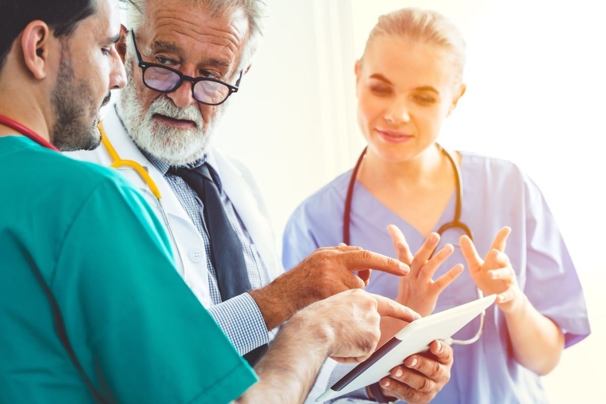 Önlük ve stetoskop giyen bir grup doktor büyük bir tablet bilgisayarın etrafında durmaktadır. Ortadaki doktor gözlüklüdür ve ekrana bakmaktadır, diğer iki doktor ise bakmaktadır. Soldaki doktor kravat takmış ve ellerini arkasında kavuşturmuş bir şekilde ayakta duruyor. Arka planda, elinde bir tablet olan bir kadın görülmektedir. Sahne, tavandan gelen parlak bir ışıkla aydınlatılmakta ve doktorların ve ellerindeki cihazın net bir şekilde görülmesini sağlamaktadır.