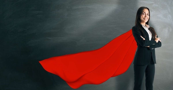 Siyah takım elbise giymiş, kollarını kavuşturmuş bir kadın kara tahtanın önünde duruyor. Yüzünde sıcak bir gülümsemeyle doğrudan kameraya bakmaktadır. Karatahtanın ortasında, koyu arka plana karşı göze çarpan parlak kırmızı bir pelerin vardır. Pelerin tahtanın üstünden aşağıya doğru sarkmakta ve aşağıya kadar uzanmaktadır. Pelerinin kumaşı pürüzsüz ve hafif dokulu, bu da ona lüks bir görünüm veriyor. Kadın koyu renk pantolon ve takım elbisesinin altına beyaz bir gömlek giymiştir. Görüntünün arka planı karanlık ve ışıklandırma yumuşak, samimi bir atmosfer yaratıyor. Kadın kendinden emin görünüyor ve varlığı sahneye hakim.