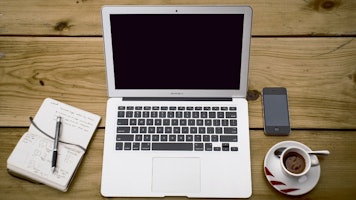 Bir dizüstü bilgisayar ve bir telefon, siyah kenarlıklı bir beyaz tahtanın önündeki ahşap bir masanın üzerinde oturuyor. Dizüstü bilgisayarın ekranı siyahtır ve telefon yakından çekilmiştir. Yanında bir kaşıkla bir fincan kahve var. Dizüstü bilgisayarda bir klavye görülüyor ve dizüstü bilgisayarın sağ tarafında bir defter açık ve üzerinde bir kalem duruyor. Kahve fincanının benzersiz bir özelliği içine çizilmiş bir yüzdür. Masa açık renklidir ve arka plan bulanıktır.