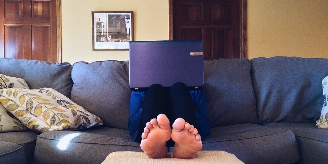 Bir kişi kanepede rahat bir pozisyonda oturmakta ve dizüstü bilgisayarı kucağında durmaktadır. Dizüstü bilgisayar mor renktedir ve beyaz bir ekranı vardır. Kişinin ayakları yakın plandan görülebiliyor ve çorap giyiyor. Odanın köşesinde çerçeve içinde bir resim görülüyor. Kanepenin üzerinde, kişinin ayaklarının yanında açık mavi renkte bir yastık var. Arka planda üzerinde mavi bir kutu bulunan ahşap bir kapı görülüyor.
