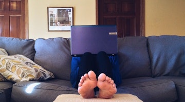 Bir kişi kanepede rahat bir pozisyonda oturmakta ve dizüstü bilgisayarı kucağında durmaktadır. Dizüstü bilgisayar mor renktedir ve beyaz bir ekranı vardır. Kişinin ayakları yakın plandan görülebiliyor ve çorap giyiyor. Odanın köşesinde çerçeve içinde bir resim görülüyor. Kanepenin üzerinde, kişinin ayaklarının yanında açık mavi renkte bir yastık var. Arka planda üzerinde mavi bir kutu bulunan ahşap bir kapı görülüyor.