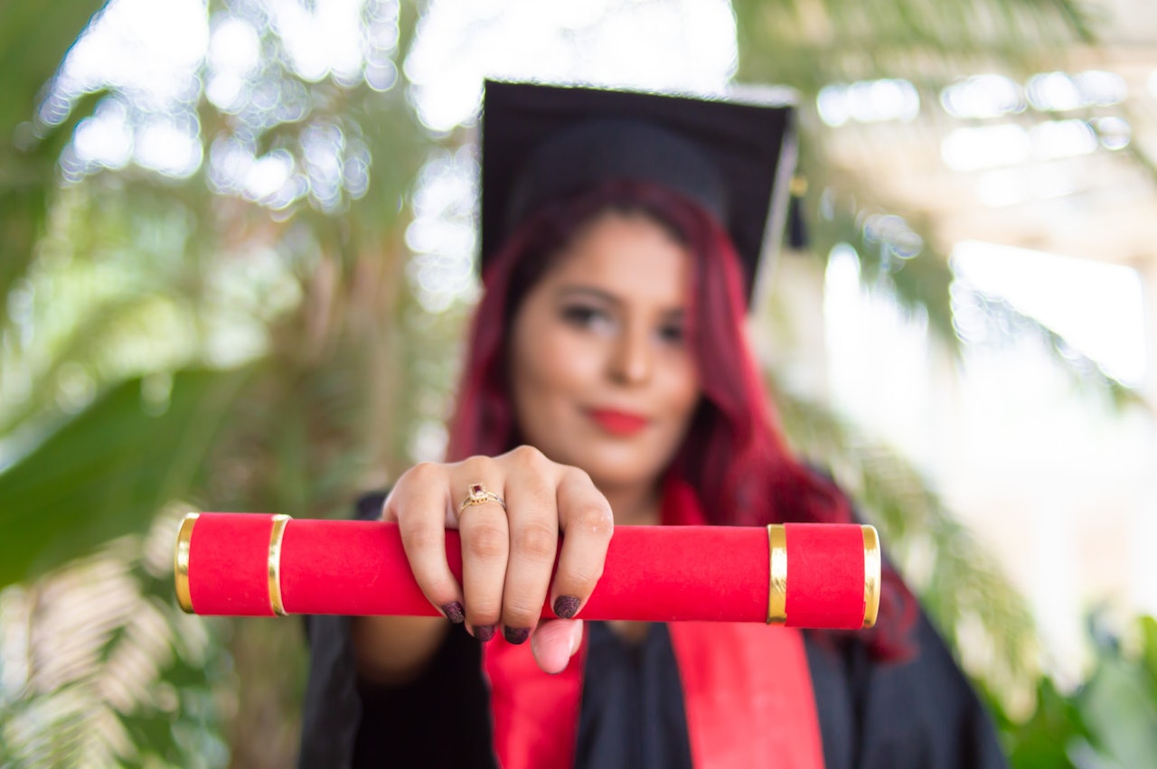 Mezuniyet cübbesi giymiş gülümseyen bir kadın, bir elinde gururla diplomasını tutuyor. Uzun, kızıl saçları var ve sarı bir püskülle süslenmiş siyah bir kep takıyor. Diğer elinde altın bir yüzük tutmaktadır ve yüzü hafifçe bulanıktır ancak sevinç ve başarı ifadesi görülebilmektedir. Büyük bir kalabalığın önünde bir sahnede durmaktadır ve güneşin ışığı üzerine vurmaktadır. Bu an önemli bir yolculuğun sonunu ve yeni bir yolculuğun başlangıcını işaret ediyor.