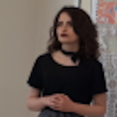 Siyah bir tişört giyen genç bir kadın profilden görünüyor. Uzun, siyah saçları yüzünden geriye doğru toplanmış ve omuzlarının üzerinde durmaktadır. Yüzünde düşünceli bir ifadeyle uzaklara bakıyor. Elleri iki yanında, kendine güvenen bir pozda duruyor. Göğsünde ve kollarında ince bir tasarım olan siyah bir gömlek giyiyor. Kumaşı yumuşak ve hafif ve vücudunu güzelce sarıyor. Cildi sıcak, doğal bir tonda parlıyor ve gözleri parlak ve uyanık. Kendinden emin ve bir amaç duygusuyla ayakta duruyor.