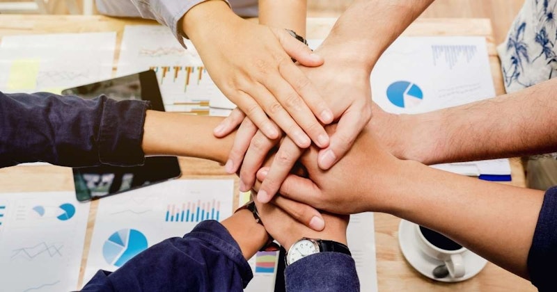 Bir grup insan birbirine yakın bir şekilde duruyor ve elleri birleşik bir şekilde kenetlenmiş. Görüntünün odak noktası, birbiriyle hafifçe örtüşen elleridir. Renk paleti sıcak ve davetkâr, maviler ve beyazlar ağırlıklı. Arka planda bir saat ve bir pasta grafiği var. Sağda, bir kişinin kolunda bir fincan kahve tutuluyor. Ön planda, bir kumaş ve bir saatin yakın çekimi görülebiliyor. Ayrıca, bir kişinin bacağı da çekimde yer alıyor. Tüm bu unsurlar bir araya gelerek görüntüdeki kişilerin ellerine odaklanan benzersiz bir an yaratıyor.