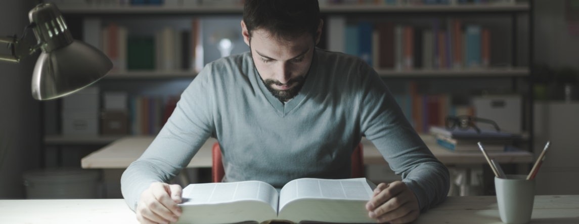 Beyaz gömlekli bir adam sandalyede oturmuş kitap okuyor. Kitabı yüzüne doğru tutuyor, sayfalar açık ve elleri kitabı alttan destekliyor. Adam gözlük takıyor ve okuduğu metne odaklandığı için yüzünde ciddi bir ifade var. Arka planda beyaz metinli siyah bir kare, metal bir nesnenin bulanık bir resmi, siyah arka planlı beyaz bir kağıt ve gölgeli beyaz bir yüzey var. Adam görüntünün merkezinde oturmakta, vücudunun üst kısmı ve yüzü belirgin bir şekilde görülmektedir.