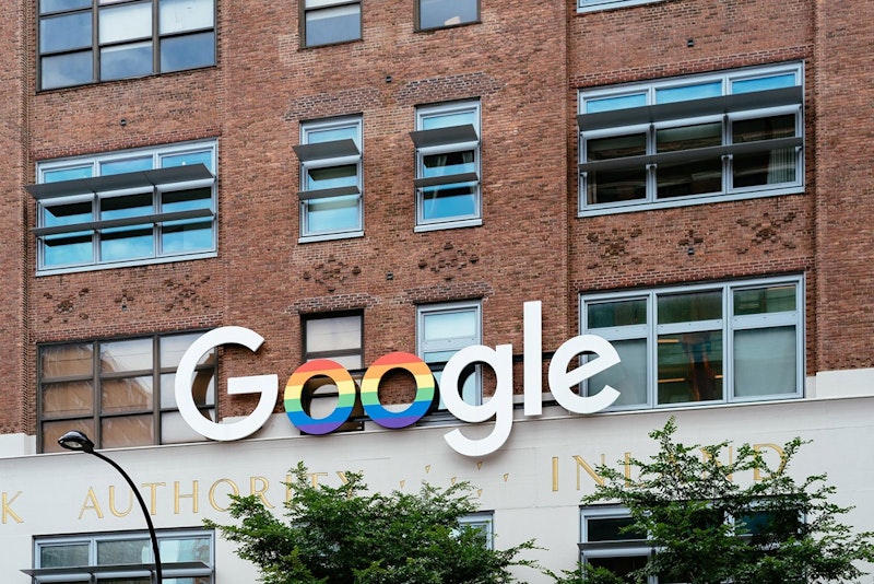 Bir binanın yakın plan görüntüsünde beyaz çerçeveli ve raflı bir pencere görülüyor. Altında gökkuşağı renginde, dairesel bir tabela asılı, üstündeki pencereyi ise beyaz bir tente örtüyor. Binanın solunda, dalları gökyüzüne uzanan bir ağaç yükseliyor. Ön planda, binanın cephesine karşı kalın harflerle yazılmış bir tabela görülüyor.