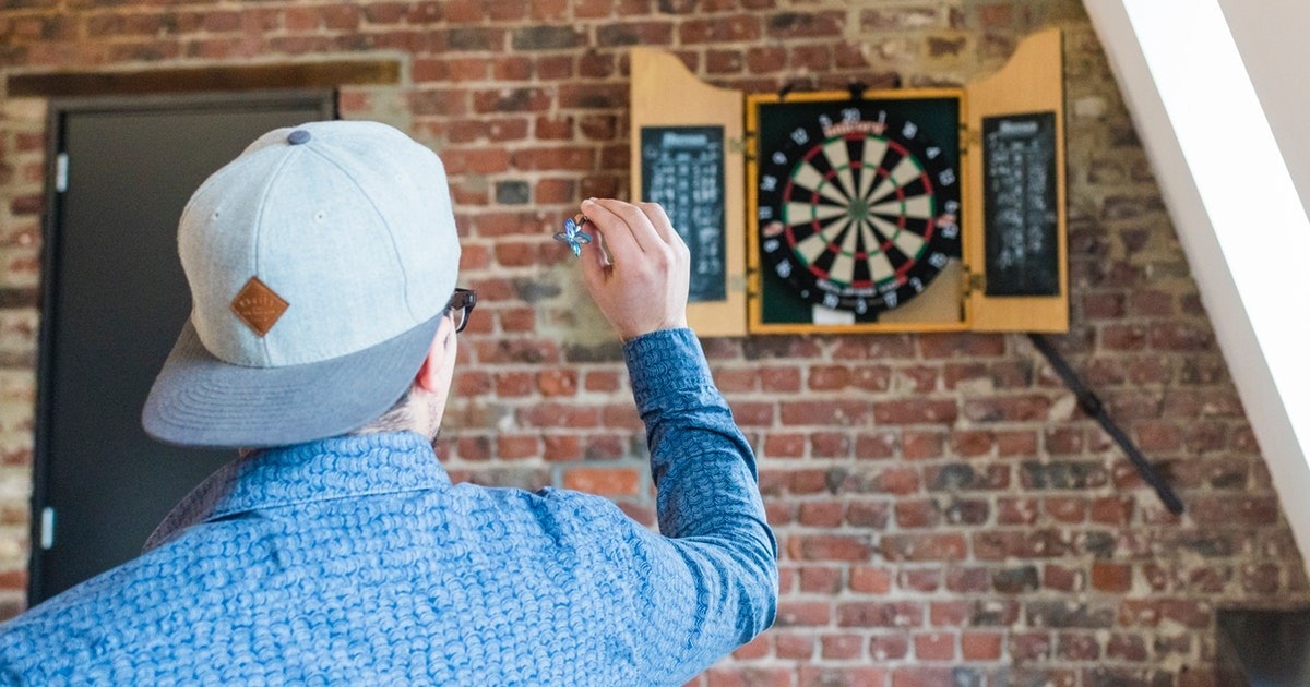 Siyah şapkalı bir adam sağ elinde bir dart, sol elinde ise mavi bir çiçek tutuyor. Arka planı bulanık olan bir dart tahtasının önünde duruyor. Dart tahtası, ortadaki koyu kahverengi bir renkte görünen elmas şeklindeki birkaç bölüme ayrılmıştır. Hedefine odaklanmış görünüyor, kolu geriye doğru çekilmiş ve atmaya hazır.