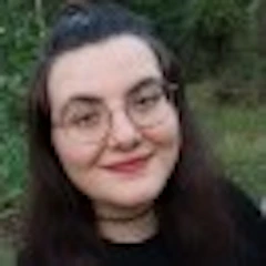 Resimdeki kadın geniş ve parlak bir gülümsemeyle gülümsüyor. Burnunun üzerinde duran ve çerçeveleri net olan bir gözlük takıyor. Gözlüklerin ardından görülebilen gözleri geniş ve ışıltılı, gerçek bir mutluluğa işaret ediyor. Yanakları hafif pembe, dudakları dolgun ve memnun bir ifadeyle kıvrılmış. Saçları koyu kahverengi ve yüzünden geriye doğru toplanmış. Resmin arka planı nötr bir renkte ve kadını resmin ana odağı haline getiriyor. Otuzlu yaşlarının başında olduğu anlaşılan kadının yüzü güneş tarafından aydınlatılmış, bu da onu rahat ve mutlu gösteriyor. Açık renkli bir bluz giyiyor ve doğrudan kameraya bakarak güzel, doğal bir portre oluşturuyor.