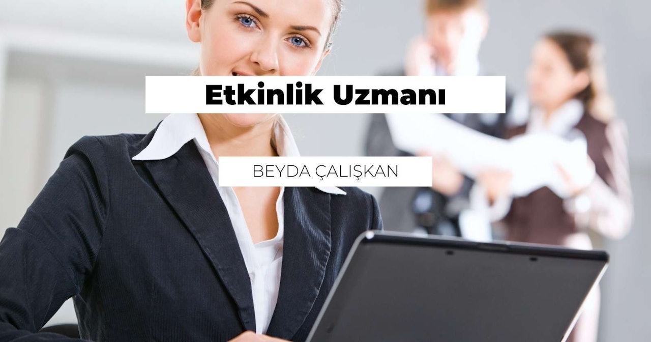 Takım elbise giyen bir kadın elinde bir dizüstü bilgisayar tutmaktadır. Yüzü yakın plandadır, burnu ve gözleri görünmektedir. Dizüstü bilgisayar açık ve ekranda siyah beyaz bir logo görülüyor. Logo bulanıktır ama kenarları seçilebilmektedir. Kadının dizüstü bilgisayarı nazikçe tutan elleri görülüyor. Dizüstü bilgisayarın klavyesi arka planda görülebiliyor. Dizüstü bilgisayar mat siyah bir yüzeye sahip ve ekran ışığı yansıtıyor.