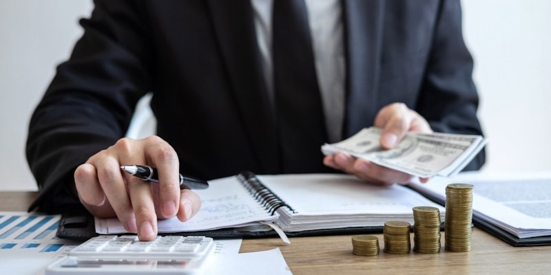 Bir kişi masada oturmuş, elindeki kalemle bir deste parayı sayıyor. Görüntünün odağı kişinin yüzü ve elleridir. Kişi beyaz bir gömlek giyiyor ve kısa siyah saçları var. Dikkatle işine konsantre olmuştur ve yüzünde ciddi bir ifade vardır. Arka planda bir klavye, bir not defteri ve bir deste altın para var. Ön planda ise bir yığın madeni para ve birkaç parça kâğıt var. Paralar altın, gümüş ve bakırdır. Kâğıt desenli bir arka plana sahip. Görüntüdeki ışık parlak ve eşittir.