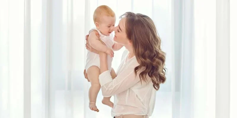 Resimde kucağında bir bebek tutan bir kadın görülüyor. Kadın beyaz bir bluz giymiş ve saçlarını geriye doğru toplamıştır. Bebeğin üzerinde pembe bir tulum var ve gözleri kapalı. Kadın bebeğe sevgi dolu bir ifadeyle bakıyor. Kollarını çok mutlu ve rahat görünen bebeğe dolamış. Çevredeki ortam nötr bir tonda ve görüntüde başka hiçbir insan ya da nesne yok. Kadının yüzü kısmen gizlendiği için bebeğin yüzü görüntünün ana odağı gibi görünüyor. Genel olarak, görüntü bir anne ile çocuğu arasındaki samimi ve hassas bir anı tasvir ediyor.
