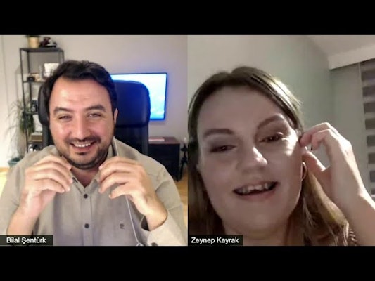 Görüntüde bir erkek ve bir kadın gülümsüyor. Adam kulaklık takmış ve bir bilgisayar ekranının önünde oturuyor. Kadın ise elini kulağına götürmüş ve doğrudan kameraya bakıyor. Görüntü çoğunlukla net, ancak bir kişinin yüzünün bulanık bir bölümü ve bir bitkinin bulanık görüntüsü var. Adam kameraya parlak bir şekilde gülümserken, kadın daha ince bir şekilde gülümsüyor. Görüntü bir erkeğin yüzünün yakın çekimidir ve erkek ve kadın aynı karede birlikte gülümsemektedir. Bu görüntü, birbirlerinin arkadaşlığından keyif alan iki insanın mükemmel bir temsilidir.