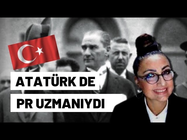 Siyah takım elbiseli ve gözlüklü bir kadın bulanık bir arka planın önünde duruyor. Saçlarını topuz yapmış ve gözlüklerini burnuna dayamış. Takım elbisesinin düğmeleri iliklenmiş ve elleri birbirine kenetlenmiş. Bakışları ileriye dönük ve ifadesi ciddi. Kendinden emin ve profesyonel görünüyor, sanki dünyayı ele geçirmeye hazır gibi. Kıyafeti ve tavırları onun başarılı bir iş kadını olduğunu gösteriyor.