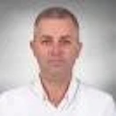 Resimdeki adam beyaz yakalı bir gömlek giyiyor. Kısa siyah saçları var ve cilt tonu hafif bronzlaşmış. Yüzü hafifçe yana eğik ve sola doğru bakıyor. Gözleri geniş ve tetikte, kaşları merakla hafifçe kalkık. Dudakları hafifçe ayrılmış ve derin düşüncelere dalmış gibi görünüyor. Temiz traşlı bir yüzü var ve çenesi hafif sivri. Yakası düzgünce ütülenmiştir ve gömleği pantolonunun içine sokulmuştur. Arka plan düz bir duvar ve görünürde hiçbir nesne yok. Dik ve kendinden emin bir şekilde duruyor, ellerini rahatça iki yanına koymuş. Ciddi bir figür, ancak ifadesi her türlü meydan okumaya hazır olduğunu gösteriyor.