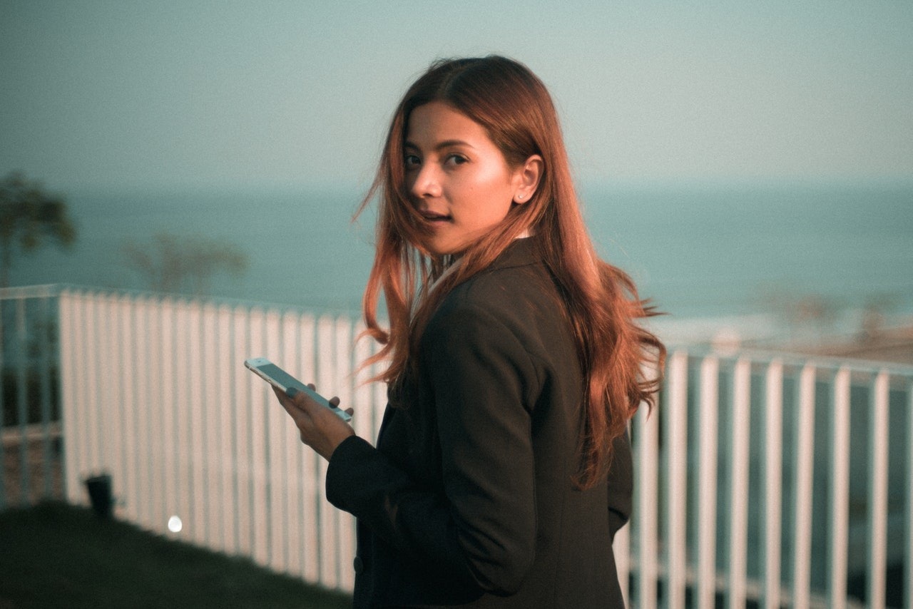 Siyah takım elbiseli bir kadın beyaz bir çitin önünde duruyor ve kulağına bir cep telefonu tutuyor. Sırtına kadar uzanan uzun, siyah saçları var. Yüzü telefonun ışığıyla aydınlanıyor ve gözleri konsantrasyon içinde kapalı. Kendinden emin ve profesyonel görünüyor. Kolunda bir saat var ve küpeleri ışıkta parlıyor. Diğer elini cebine sokmuş. Arka planda parlak, neşeli bir gökyüzü var ve güneş onun üzerinde parlıyor.