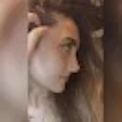 Bu bir kadının yüzünün yakın çekim fotoğrafı. Sıcak, pembe bir alt tona sahip açık tenli görünüyor. Gözleri badem şeklinde ve kaşları ince ve zarif bir şekilde kemerli. Dudakları dolgun ve dolgun ve burnu küçük, düğme benzeri bir şekle sahip. Sağ yanağında, burnunun yanında bir güzellik izi vardır. İfadesi nazik ve huzurlu, bir parça da memnuniyet içeriyor. Çenesinde küçük bir gamzesi vardır ve saçları yüzünden geriye doğru taranmıştır. Öne çıkan ve parlayan çok doğal, klasik bir güzelliği var.