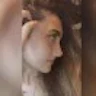 Bu bir kadının yüzünün yakın çekim fotoğrafı. Sıcak, pembe bir alt tona sahip açık tenli görünüyor. Gözleri badem şeklinde ve kaşları ince ve zarif bir şekilde kemerli. Dudakları dolgun ve dolgun ve burnu küçük, düğme benzeri bir şekle sahip. Sağ yanağında, burnunun yanında bir güzellik izi vardır. İfadesi nazik ve huzurlu, bir parça da memnuniyet içeriyor. Çenesinde küçük bir gamzesi vardır ve saçları yüzünden geriye doğru taranmıştır. Öne çıkan ve parlayan çok doğal, klasik bir güzelliği var.