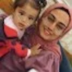 Bu, kucağında küçük bir çocuk taşıyan bir kadının görüntüsüdür. Kadın açık renkli bir elbise giymiş ve saçlarını geriye doğru toplamış, yüzü nazik bir ifadeyle çocuğa bakıyor. Çocuk desenli bir gömlek giymiş, açık renkli saçları ve koyu renk gözleri var, yüzü kadının göğsüne yaslanmış. Kadının kolları çocuğa sarılmış, bir güvenlik ve sevgi hissi veriyor. Arka plan bulanık, ağaçlar ve çimenlerle dolu bir park ortamını andırıyor. Görüntü, anne ve çocuk arasındaki güçlü bağı aktaran sıcaklık ve şefkat dolu.