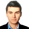 Resimdeki adam gri bir takım elbise giyiyor ve altında beyaz bir gömlek var. Düzgün, kısa siyah saçları ve temiz traşlı bir yüzü var. Parke zeminli, beyaz duvarlı ve arkasında pencere olan bir odada duruyor. Kendinden emin görünüyor, bakışları doğrudan ve duruşu dik. Elleri önünde kenetlenmiş ve sol bileğinde bir saat takılı. Kırklı yaşlarının ortasında ve profesyonelce giyinmiş görünüyor. İyi giyimli bir iş adamının mükemmel bir örneği.