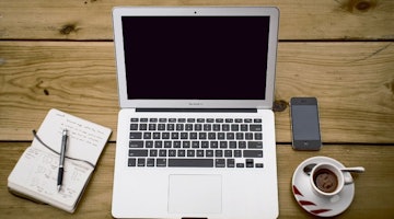 Bir dizüstü bilgisayar ve bir telefon ahşap bir masanın üzerine yerleştirilmiştir. Dizüstü bilgisayar siyah ekranlı ve beyaz klavyeli beyaz bir bilgisayar, telefon ise siyah ekranlı ve siyah bir telefon. Dizüstü bilgisayarın yanında siyah ekranlı ve siyah kenarlıklı beyaz bir tablet var. Telefonun yanında kaşıklı bir fincan kahve, masanın diğer ucunda ise siyah kenarlıklı beyaz bir tahta yer alıyor. Dizüstü bilgisayarın yanındaki defterin üzerinde bir kalem durmaktadır. Kahve fincanının üzerine bir yüz çizilmiştir.