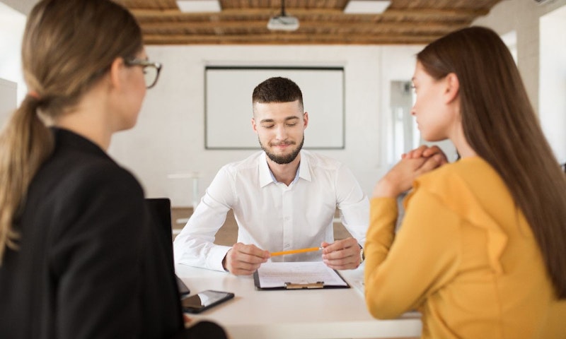 Kısa saçlı ve sakallı bir adam, masada oturan bir kadının önünde kalem tutmaktadır. Kadın beyaz bir bluz giymiş ve ellerini birleştirmiş. Adam gri bir gömlek giymiş ve yüzünde ciddi bir ifade var. Önlerindeki masada içinde bir kağıt parçası olan bir telefon var. Görüntünün arka planında, damarları yakından çekilmiş bir ahşap var. Kadının at kuyruğu var ve tüm dikkatini adama veriyor. Dikkatle bir şey tartışıyor gibi görünüyorlar.