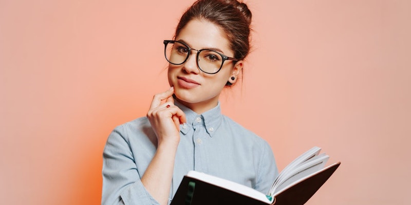 Gözlüklü bir kadın elinde bir kitap tutmaktadır. Gözleri kitaba odaklanmış ve kolları hafifçe içe doğru bükülmüş. Kitabın üzerinde hafif bir desen bulunan beyaz bir kapağı var ve sayfalar hafifçe açılmış. Kadının koyu renk saçları var ve yüzü kitap tarafından gizlenmiş. Arka plan, üzerinde birkaç kırışıklık ve çizgi bulunan açık renkli bir yüzeydir. Görüntü yakın çekimdir ve kadının parmakları kitabı kavrarken görülebilmektedir. Kadın düşünceli ve kitaba odaklanmış görünüyor.