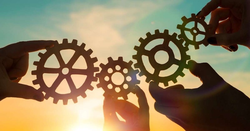 Bir grup el, çeşitli boyut ve şekillerde çeşitli dişliler tutuyor. Bir ele odaklanan izleyici, başparmak ile işaret parmağı arasında tutulan bir dişli görebiliyor. Dişli koyu gümüş rengindedir ve üzerinde birkaç çentik ve kabarık bir desen bulunan kavisli bir yüze sahiptir. İlk elin arkasında, izleyici daha geniş bir çevreye ve düz bir yüze sahip farklı bir dişli tutan başka bir el görebilir. Arka plan açık gri bir renk tonuna sahip ve eller ile dişliler görüntünün odak noktasını oluşturuyor. Tüm eller birbirine yakın tutularak güçlü ve birleşik bir kompozisyon yaratılmış. Görüntü, her bir elin kolektif çabada önemli bir rol oynadığı bir ekip çalışması ve işbirliği duygusu aktarıyor.