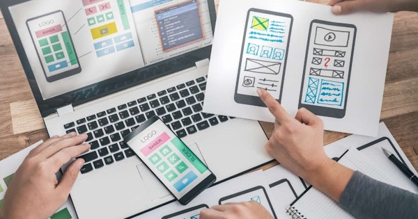 Bir kişi dizüstü bilgisayar ve cep telefonunu birlikte kullanıyor. Kişi bir kağıt parçası üzerindeki bir çizimi işaret ediyor ve diğer eli dizüstü bilgisayarın klavyesinde. Bir kalemin yakın çekimi var ve kişinin eli de bir parça kağıt tutuyor. Çizim bir tel kafes veya bir web sayfası gibi görünüyor. Kişi çizime odaklanmış gibi görünüyor ve ifadesi düşünceli.