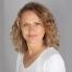 Resimdeki kadının ortadan ayrılmış ve hafifçe geriye taranmış uzun, düz saçları var. Kolları kıvrılmış ve üst düğmeleri açık bırakılmış beyaz, yakalı bir gömlek giyiyor. Doğal, sıcak bir gülümsemesi var ve bakışları uzaklara yönelmiş. Yüzünü yumuşak ve parlak, omuzlarına dökülen saçları çerçeveliyor. Teni açık ten rengi, gözleri ise koyu kahverengidir. Kıyafeti sade ve gündelik, duruşu rahat ve kendinden emin. Bulunduğu ortamda sakin, memnun ve rahat görünüyor. Güzel bir manzara ve varlığı huzur ve dinginlik hissi yayıyor.