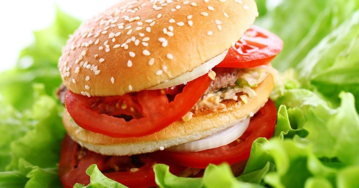 Bu görüntü domatesli ve marullu bir hamburgerin yakın çekimidir. Hamburger açık gri bir arka plan üzerine yerleştirilmiştir ve domates dilimleri parlak kırmızıdır. Marul yaprakları parlak yeşil renkte ve hamburgerin etrafına dizilmiş. Burgerin üzerine kalın bir dilim sarı peynir yerleştirilmiştir. Hamburger ikiye bölünerek sandviçin içeriği ortaya çıkarılmıştır. Domates dilimleri dairesel bir şekilde dizilir ve burgerin içine iki turşu yerleştirilir. Burgerin üzerine sandviçe lezzetli bir tat veren açık kahverengi bir sos gezdirilir. Marul yaprakları burgere gevrek bir doku katmaktadır. Hamburgerin genel sunumu görsel olarak çekicidir ve onu lezzetli bir yemek haline getirir.