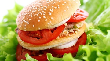 Bu görüntü domatesli ve marullu bir hamburgerin yakın çekimidir. Hamburger açık gri bir arka plan üzerine yerleştirilmiştir ve domates dilimleri parlak kırmızıdır. Marul yaprakları parlak yeşil renkte ve hamburgerin etrafına dizilmiş. Burgerin üzerine kalın bir dilim sarı peynir yerleştirilmiştir. Hamburger ikiye bölünerek sandviçin içeriği ortaya çıkarılmıştır. Domates dilimleri dairesel bir şekilde dizilir ve burgerin içine iki turşu yerleştirilir. Burgerin üzerine sandviçe lezzetli bir tat veren açık kahverengi bir sos gezdirilir. Marul yaprakları burgere gevrek bir doku katmaktadır. Hamburgerin genel sunumu görsel olarak çekicidir ve onu lezzetli bir yemek haline getirir.