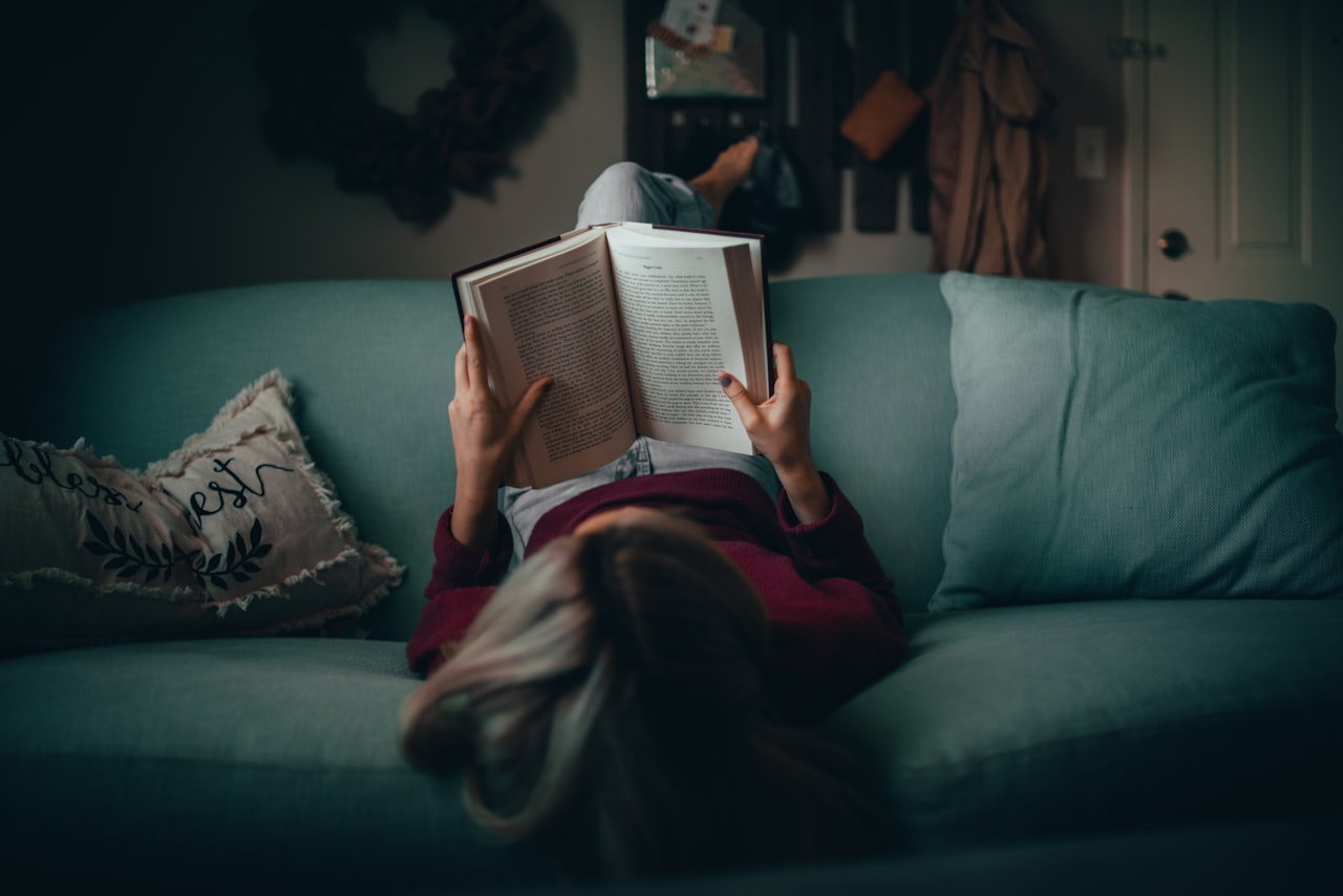 Bir kadın kanepede rahat bir pozisyonda uzanmış, kitap okuyor. Mavi kot pantolon ve beyaz bir tişört giymiş olan kadının başı, üzerinde püsküller bulunan renkli bir yastıkla desteklenmiş. Sağ eli açık kitabı yüzünün önünde tutuyor ve sol kolu kanepenin kol dayanağına dayanıyor. Arka plan biraz bulanık, ancak arkasında kapı tokmağı olan beyaz bir kapı görülebiliyor.