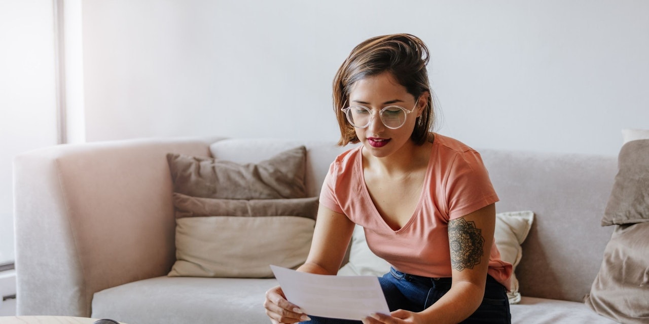 A woman is sitting on a couch and reading a newspaper. She is wearing glasses and has a tattoo on her left arm. Her lips are painted red and she is wearing a pink shirt. There is a close-up of her face, showing her glasses, and her eyes are focused intently on the paper. Her hair is pulled back in a neat bun, and her expression is focused and concentrated. The newspaper she is reading is held in her hands, and she is flipping through the pages.