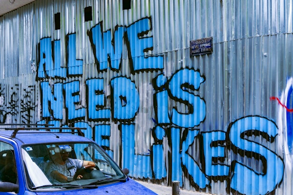 Mavi arabalı bir adam, grafitiyle kaplı metal bir duvarın önünden geçmektedir. Duvar mavi, beyaz ve siyah grafitilerle boyanmış ve duvarın tepesine siyah bir kare yapıştırılmıştır. Arabanın yanına yeşil etiketli metal bir çöp kutusu park edilmiş ve duvarda beyaz yazılı bir tabela var. Ön planda bir pencerenin yakın çekimi görülüyor. Adam güneş gözlüğü ve beyaz bir gömlek giymiştir ve dümdüz karşıya bakmaktadır. Araba sabit bir hızla ilerliyor ve güneş pırıl pırıl parlıyor.